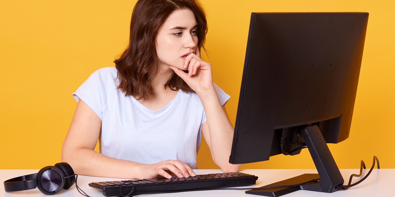 Studio Shot Of Pleasant Looking Student With Concentrated Look, Wears Casual Outfit, Poses Against Yellow Background. Beautiful Woman Working Hard, Earns Money Online, Keeps Hand Under Chin.